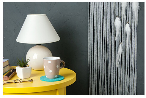 yellow bedside table with white lamp and decorative items on top, all against dark grey wall 